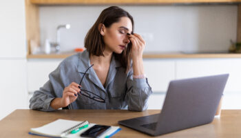 A young woman in a gray shirt sits at a desk with a laptop, rubbing the bridge of her nose while holding her glasses, looking fatigued. Signs of depression can include persistent exhaustion, trouble focusing, and feeling overwhelmed.