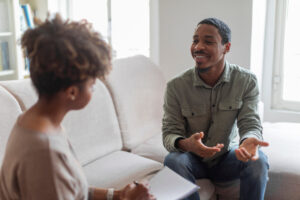 Cheerful positive handsome young african american guy in casual outfit sitting on couch at cozy counselor office, having conversation with therapist black woman, sharing good because he decided to start seeing a therapist