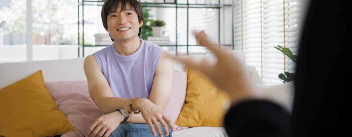 A non-binary Asian individual sits comfortably on a couch during a face-to-face session with an LGBTQ+ affirming therapist, in a bright, welcoming room with natural light and homey decor.