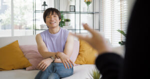 A non-binary Asian individual sits comfortably on a couch during a face-to-face session with an LGBTQ+ affirming therapist, in a bright, welcoming room with natural light and homey decor.