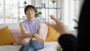 A non-binary Asian individual sits comfortably on a couch during a face-to-face session with an LGBTQ+ affirming therapist, in a bright, welcoming room with natural light and homey decor.