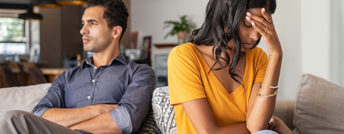 Young couple sitting on couch after an argument. Sad woman sitting with hand on head after quarrel with boyfriend at home. Angry couple ignoring each other on the sofa, having relationship troubles.