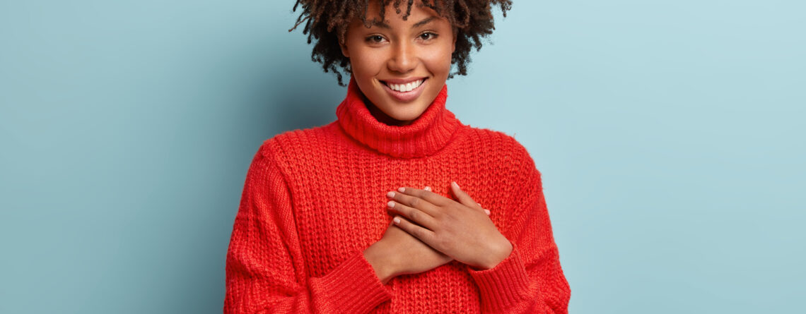 Photo of emotional dark skinned female touches heart with both palms, demonstrates thankful gesture, has good natured expression, expresses great sympathy, gratitude with honesty, feels love